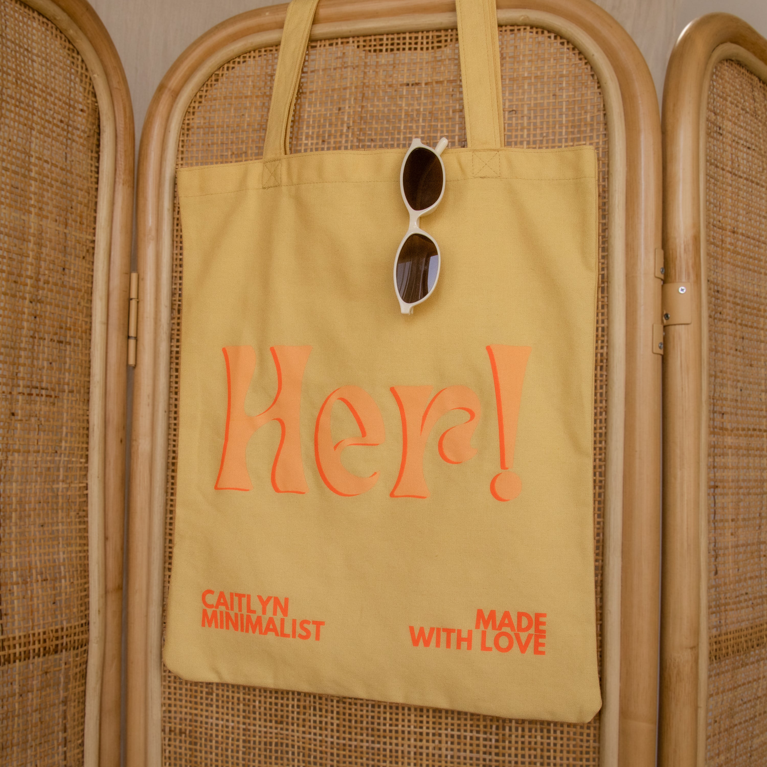 Brighten your day with the Free Gift of Choice—a yellow tote bag featuring "Her!" in orange text and sunglasses detail, making it a perfect pick for tote bag lovers or as a cheerful free gift.