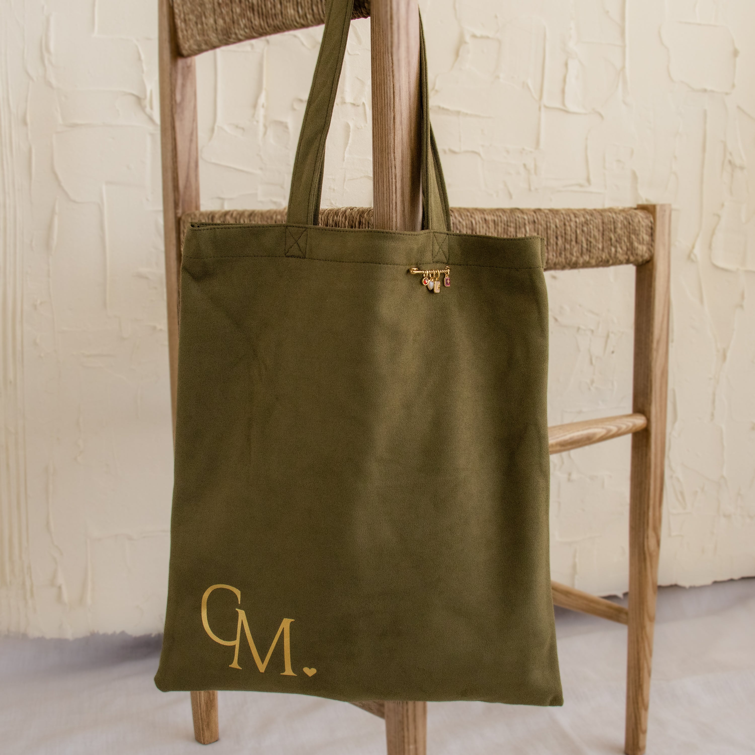 Olive green "Free Gift of Choice" tote bag with CM. in gold lettering, hanging on a wooden chair with woven seat—a stylish addition to your tote collection.