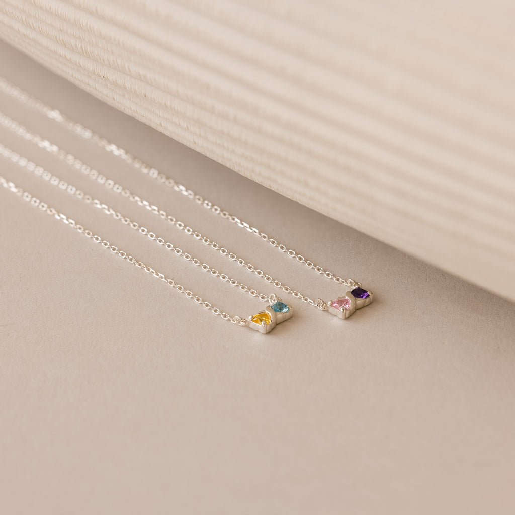 The Toi et Moi Infinity Heart Birthstone Necklace, a delicate silver piece featuring a colorful gemstone pendant, is displayed on a neutral background.