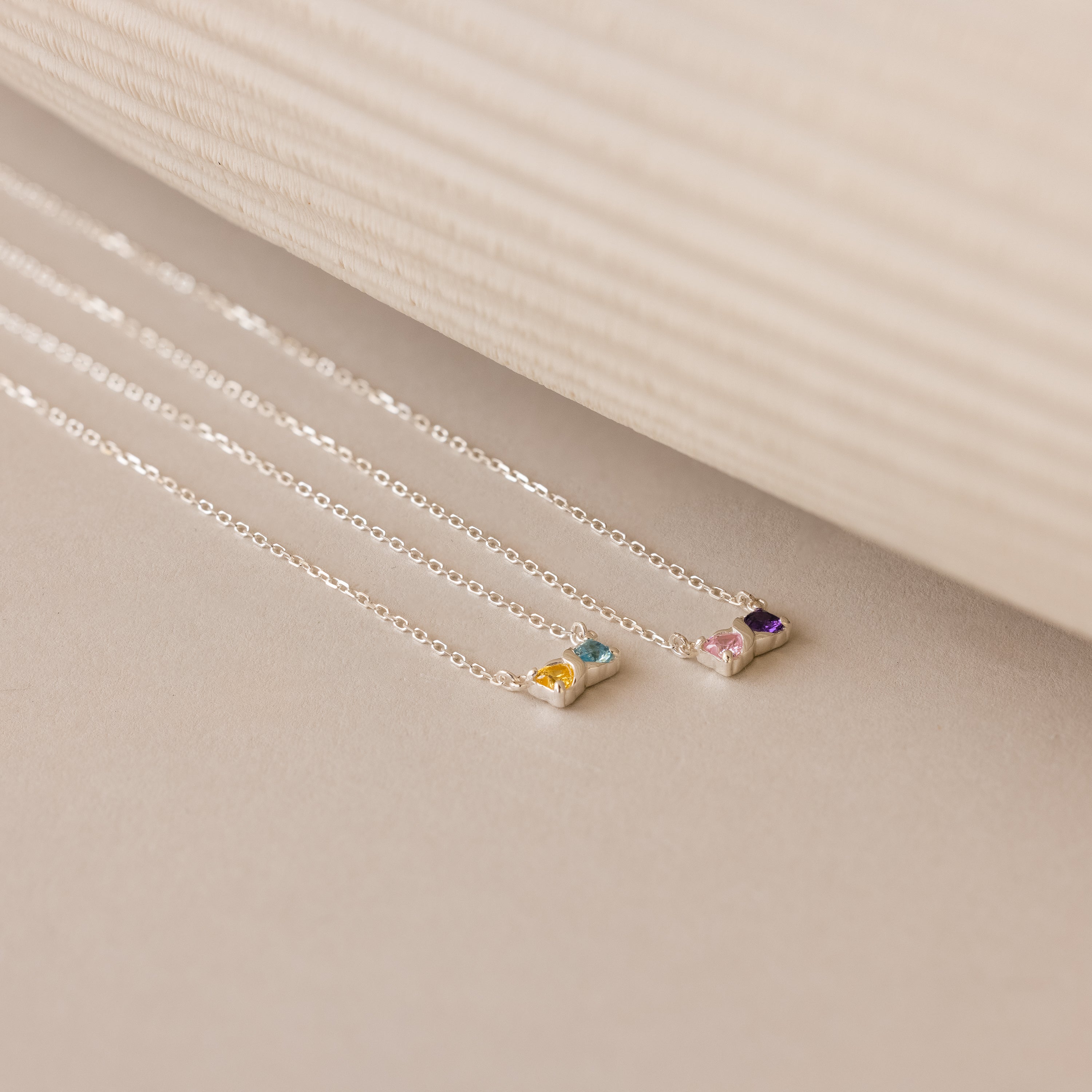The Toi et Moi Infinity Heart Birthstone Necklace, a delicate silver piece featuring a colorful gemstone pendant, is displayed on a neutral background.