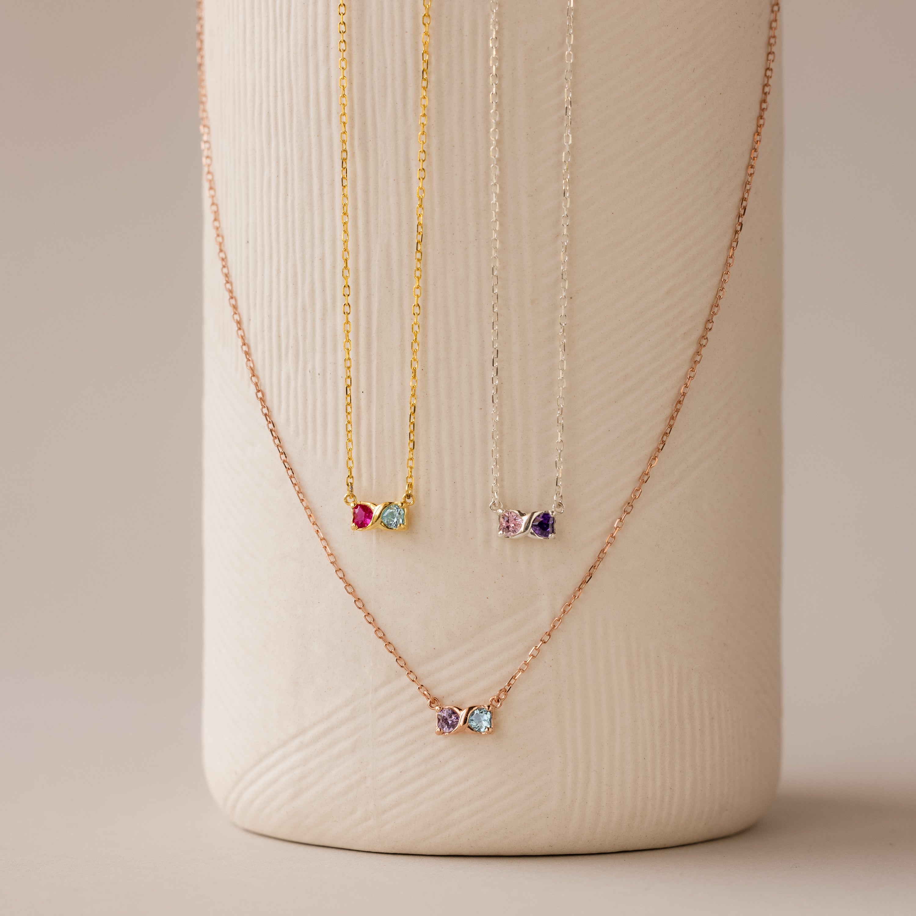 The Toi et Moi Infinity Heart Birthstone Necklaces, featuring colorful gemstone pendants, are displayed on a textured white vase—perfect for adding a personal touch to any look.