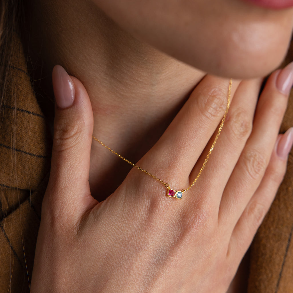 A woman touches her neck, wearing the Toi et Moi Infinity Heart Birthstone Necklace—a delicate gold chain featuring a small red gemstone pendant for an elegant, personalized look.