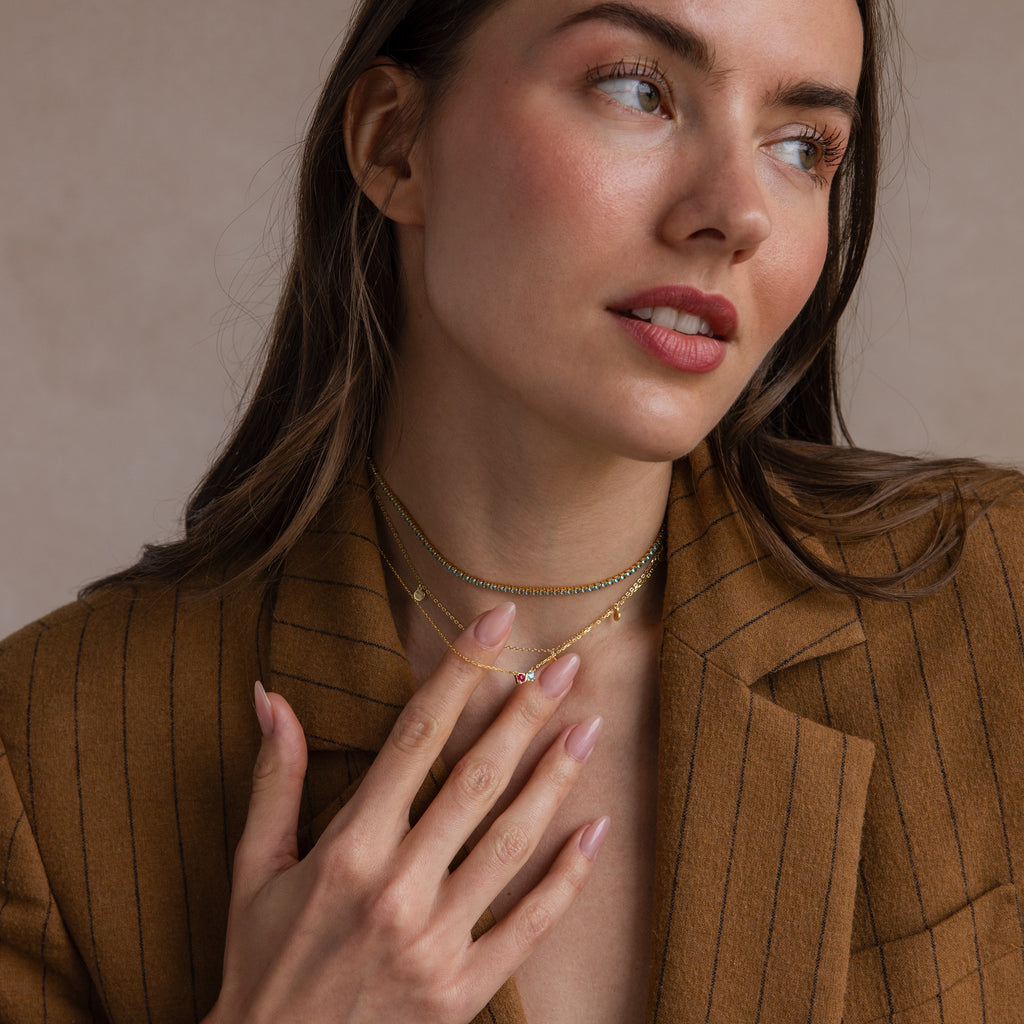 A woman in a brown pinstripe blazer touches her Toi et Moi Infinity Heart Birthstone Necklace and gold chains, gazing softly to the side.
