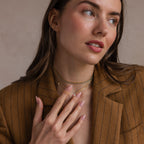 A woman in a brown pinstripe blazer touches her Toi et Moi Infinity Heart Birthstone Necklace and gold chains, gazing softly to the side.