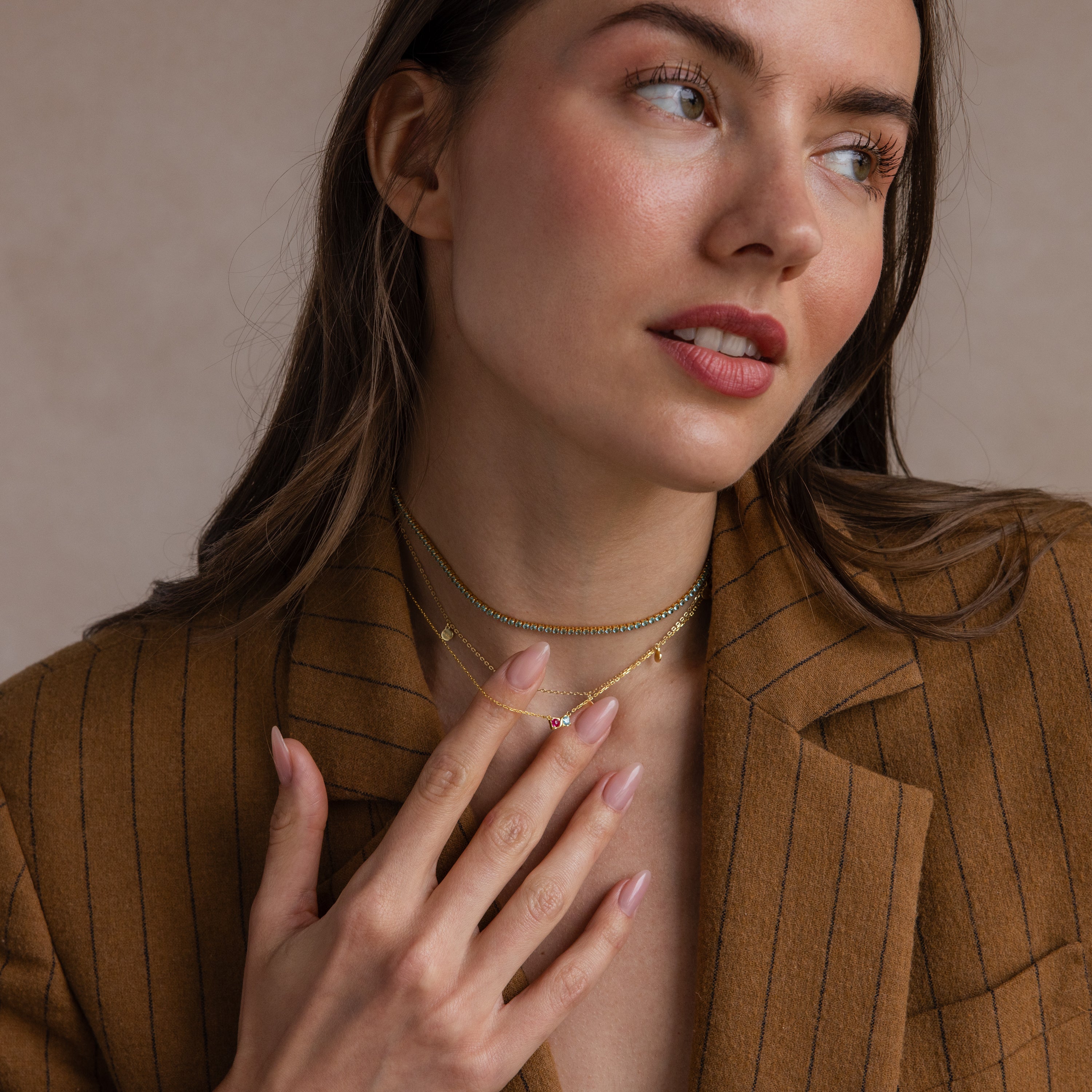 A woman in a brown pinstripe blazer touches her Toi et Moi Infinity Heart Birthstone Necklace and gold chains, gazing softly to the side.