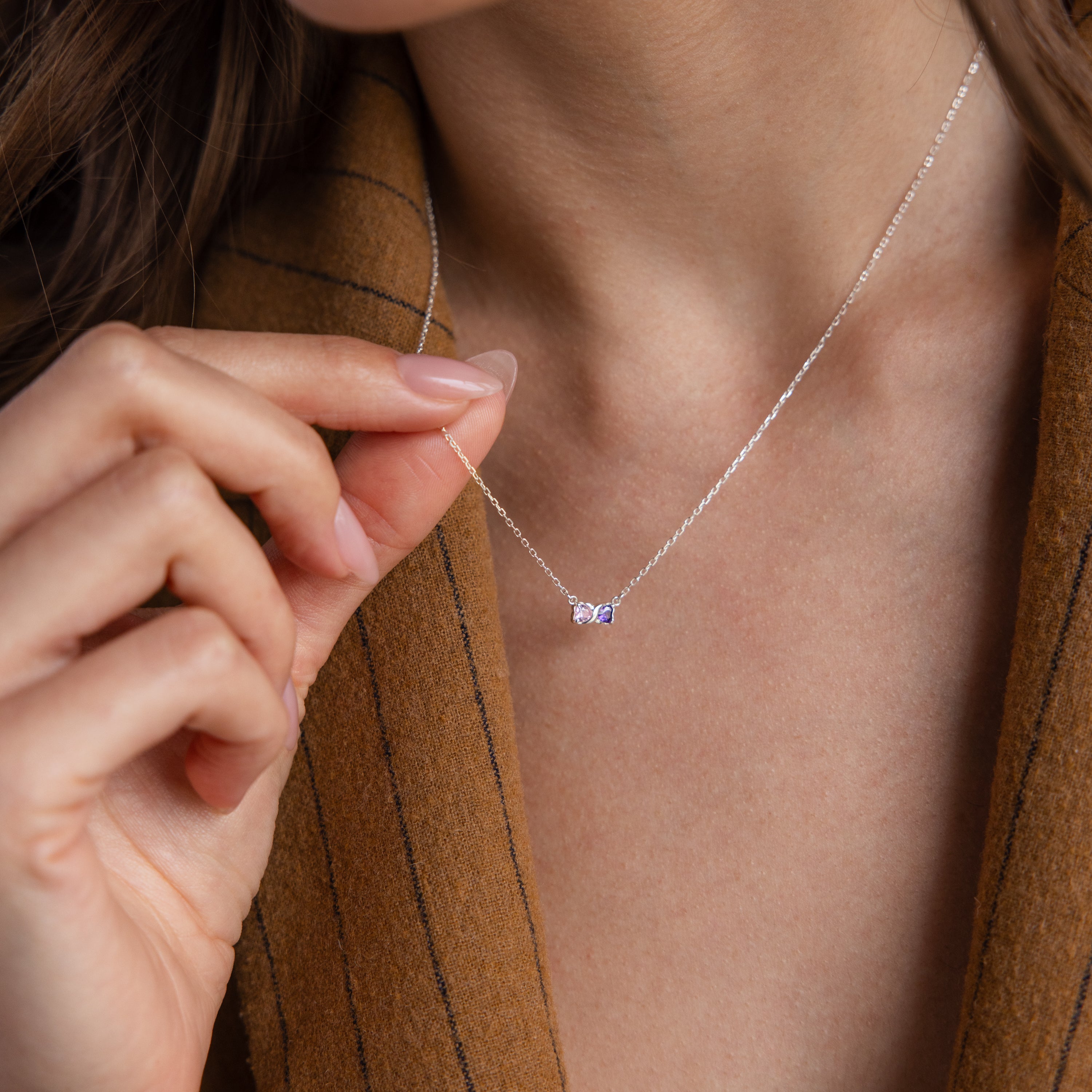 A woman wearing a brown pinstripe blazer holds the Toi et Moi Infinity Heart Birthstone Necklace, featuring a delicate pendant.