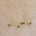 Five Toi et Moi Infinity Heart Birthstone Necklaces, each adorned with two colorful gemstones, are displayed on a beige marble surface—perfect personalized jewelry for birthstone lovers.