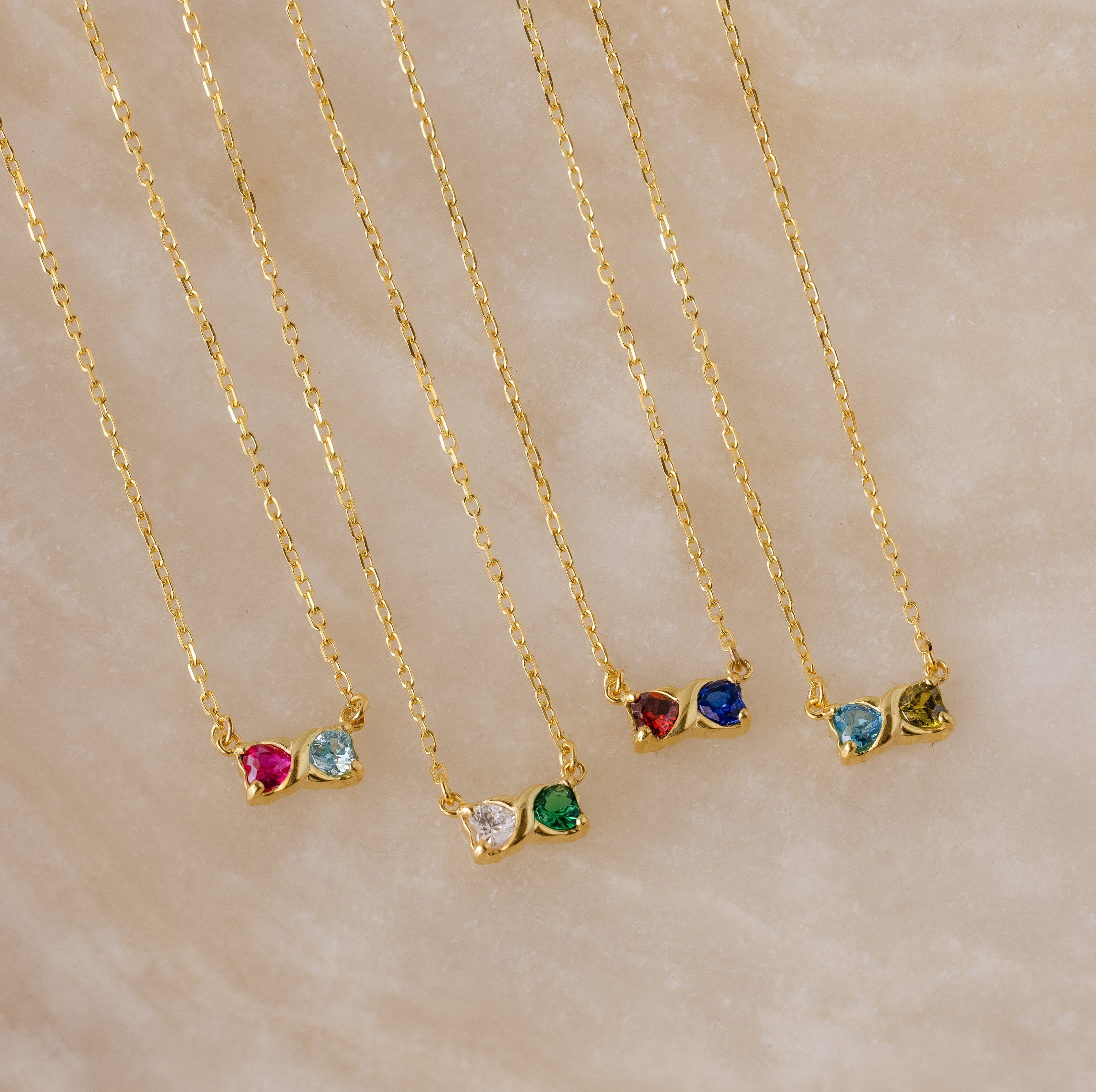 Five Toi et Moi Infinity Heart Birthstone Necklaces, each adorned with two colorful gemstones, are displayed on a beige marble surface—perfect personalized jewelry for birthstone lovers.