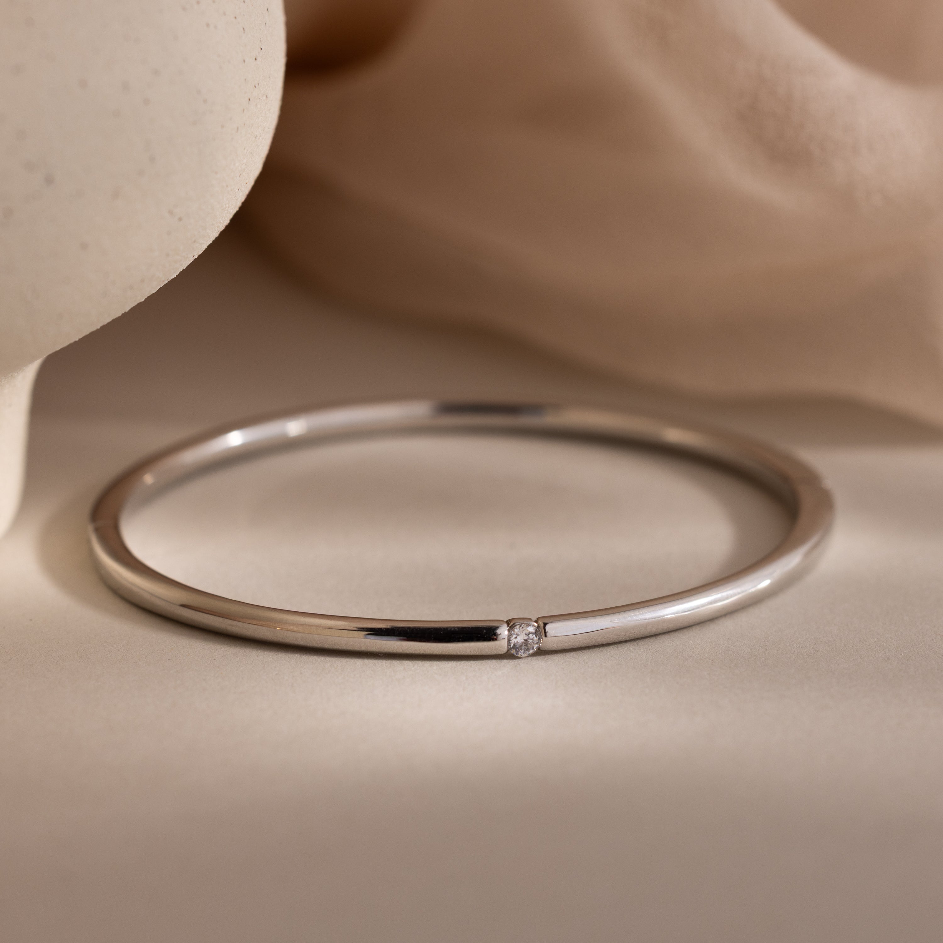The Skinny Diamond Bangle in White Gold is a minimalist bracelet with a small diamond accent, elegantly showcased on a soft beige fabric background.