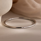 The Skinny Diamond Bangle in White Gold is a minimalist bracelet with a small diamond accent, elegantly showcased on a soft beige fabric background.