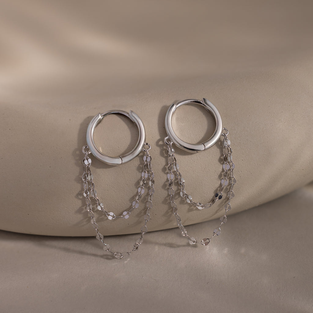 The Duo Mirror Chain Huggies in Sterling Silver feature two delicate chain strands and are shown on a smooth beige surface—ideal for those who love modern jewelry.