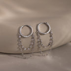 The Duo Mirror Chain Huggies in Sterling Silver feature two delicate chain strands and are shown on a smooth beige surface—ideal for those who love modern jewelry.