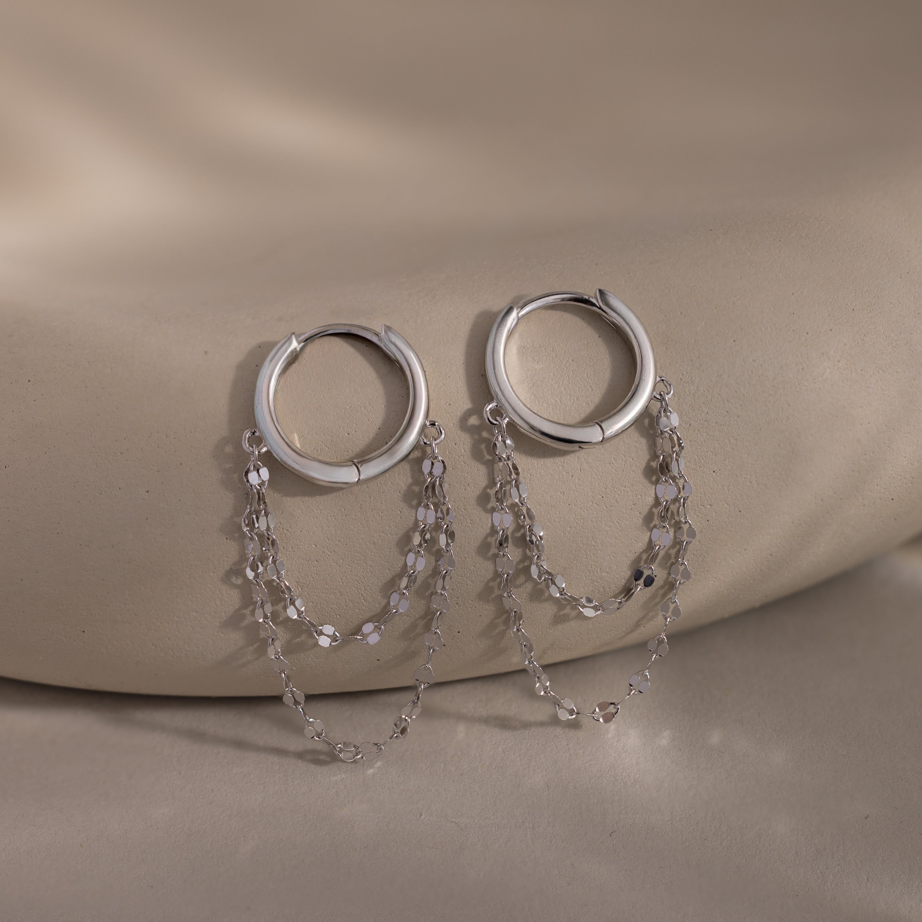 The Duo Mirror Chain Huggies in Sterling Silver feature two delicate chain strands and are shown on a smooth beige surface—ideal for those who love modern jewelry.