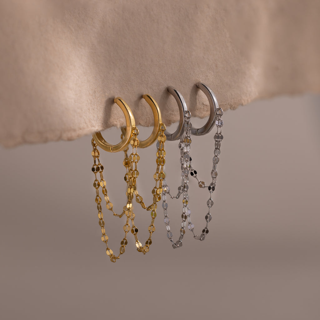 The Duo Mirror Chain Huggies, featuring double-layered chains in both gold and silver, hang elegantly on a textured beige surface—perfect for modern jewelry lovers.