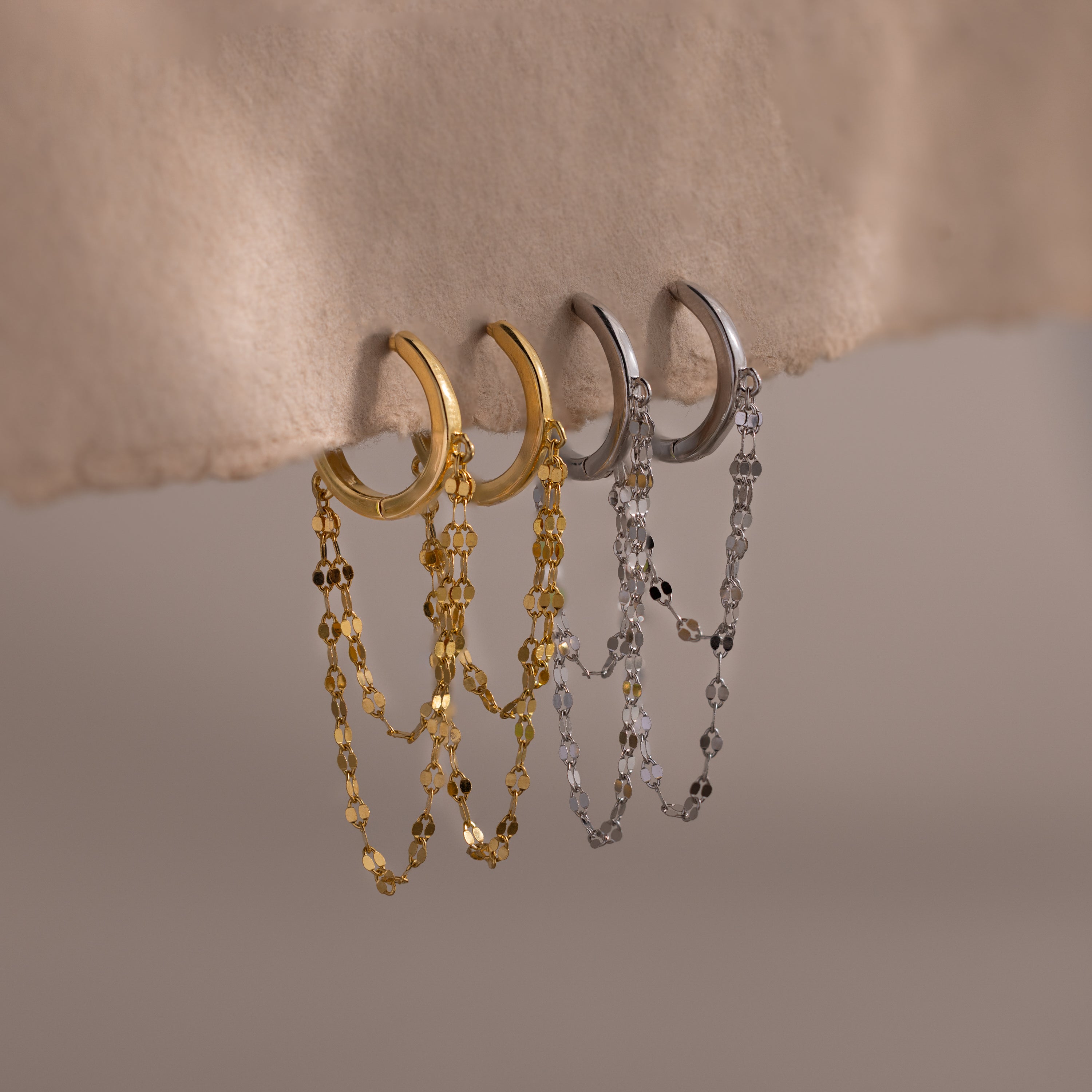 The Duo Mirror Chain Huggies, featuring double-layered chains in both gold and silver, hang elegantly on a textured beige surface—perfect for modern jewelry lovers.