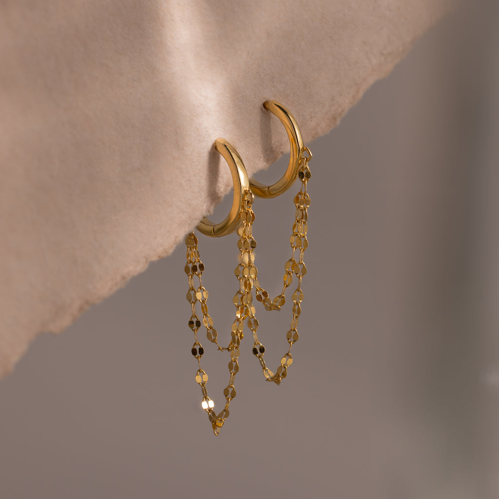 Duo Mirror Chain Huggies in 18K Gold, featuring double layered chain details, offer a modern jewelry touch and are displayed on a soft beige fabric background.