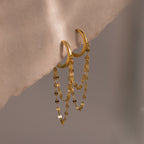 Duo Mirror Chain Huggies in 18K Gold, featuring double layered chain details, offer a modern jewelry touch and are displayed on a soft beige fabric background.