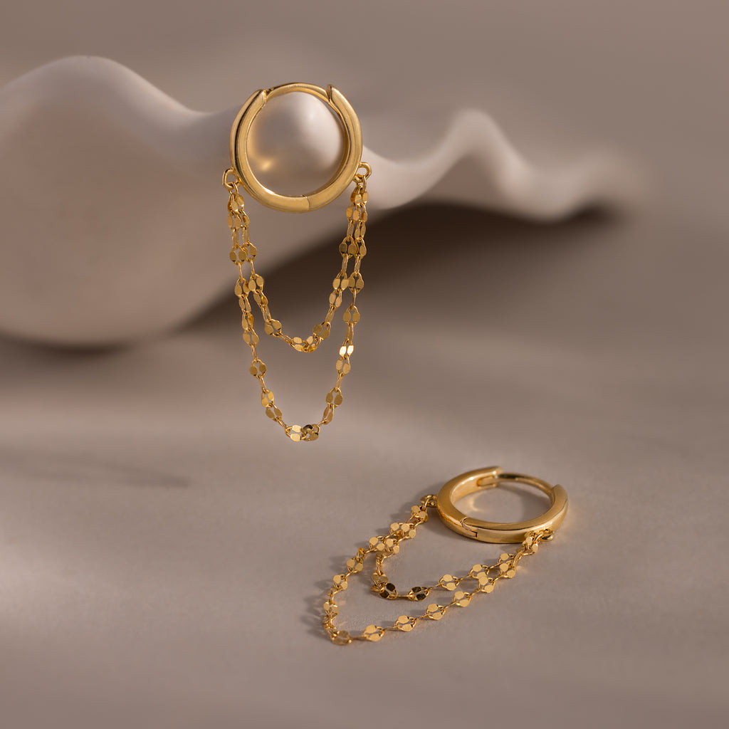 Duo Mirror Chain Huggies in 18K Gold—one displayed upright on a sculpted white surface, the other lying flat—capture the layered earrings style for fans of modern jewelry.