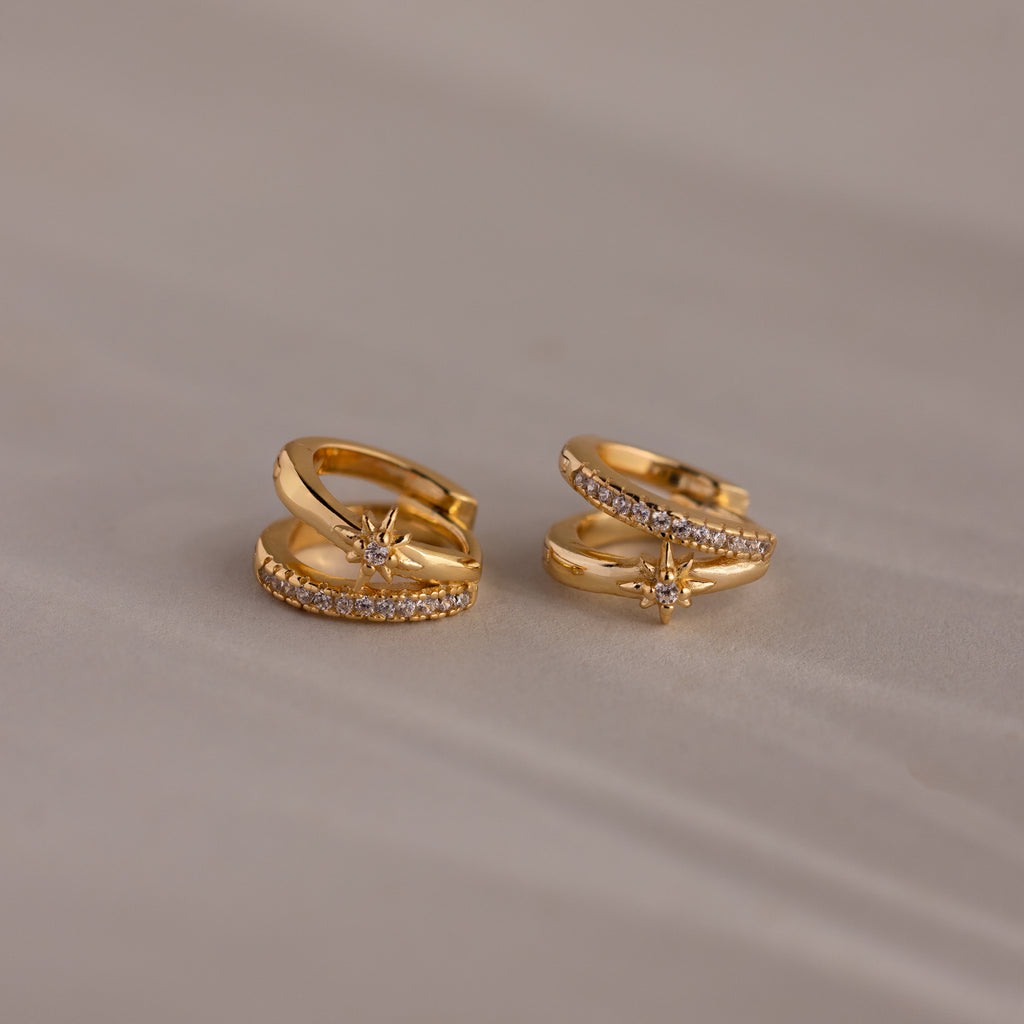 The Starburst Pave Duo Huggies, gold hoops featuring a star-shaped design with small clear stones, are shown on a light beige surface.
