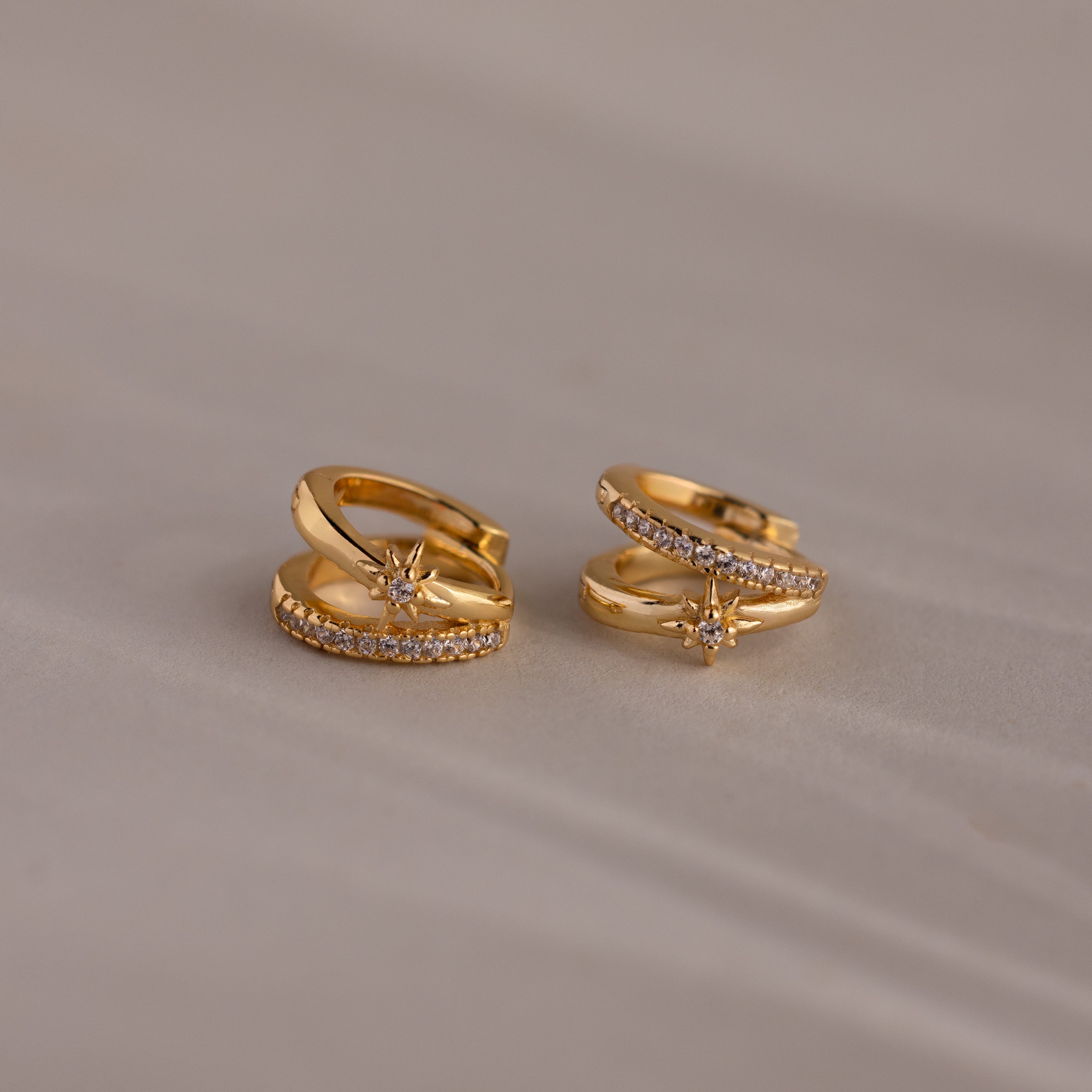 The Starburst Pave Duo Huggies, gold hoops featuring a star-shaped design with small clear stones, are shown on a light beige surface.