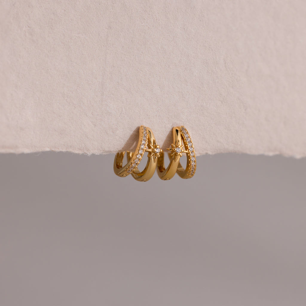 The Starburst Pave Duo Huggies feature two gold hoops—one with sparkling crystals, the other with a star charm—beautifully displayed on textured paper.