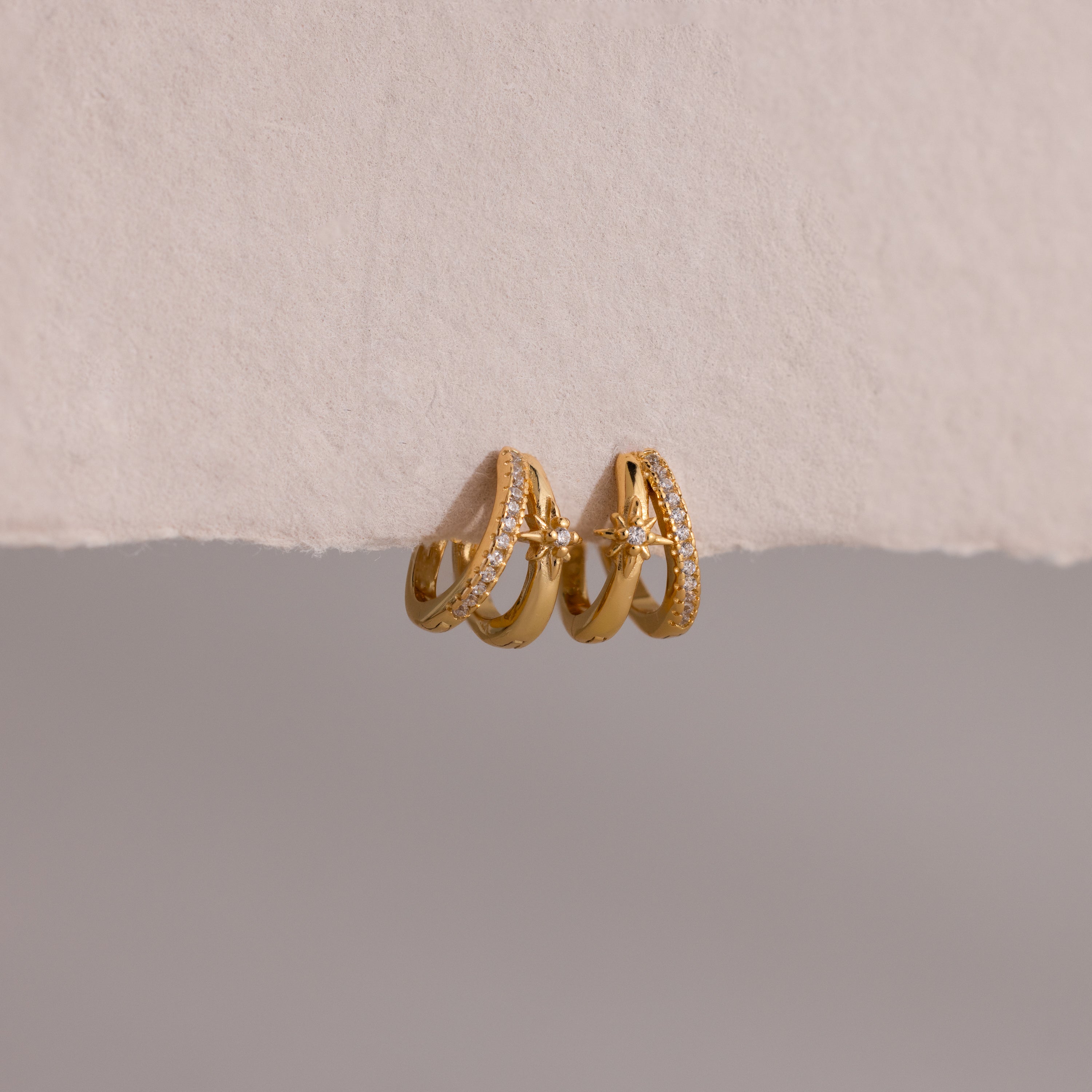 The Starburst Pave Duo Huggies feature two gold hoops—one with sparkling crystals, the other with a star charm—beautifully displayed on textured paper.