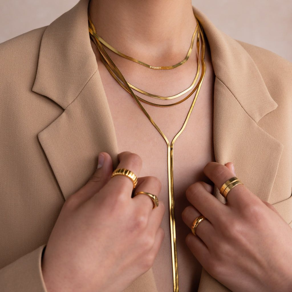 Person wearing the Long Y Herringbone Chain Necklace in 18K Gold layered with other gold necklaces and rings, dressed in a beige blazer with hands near the collar.