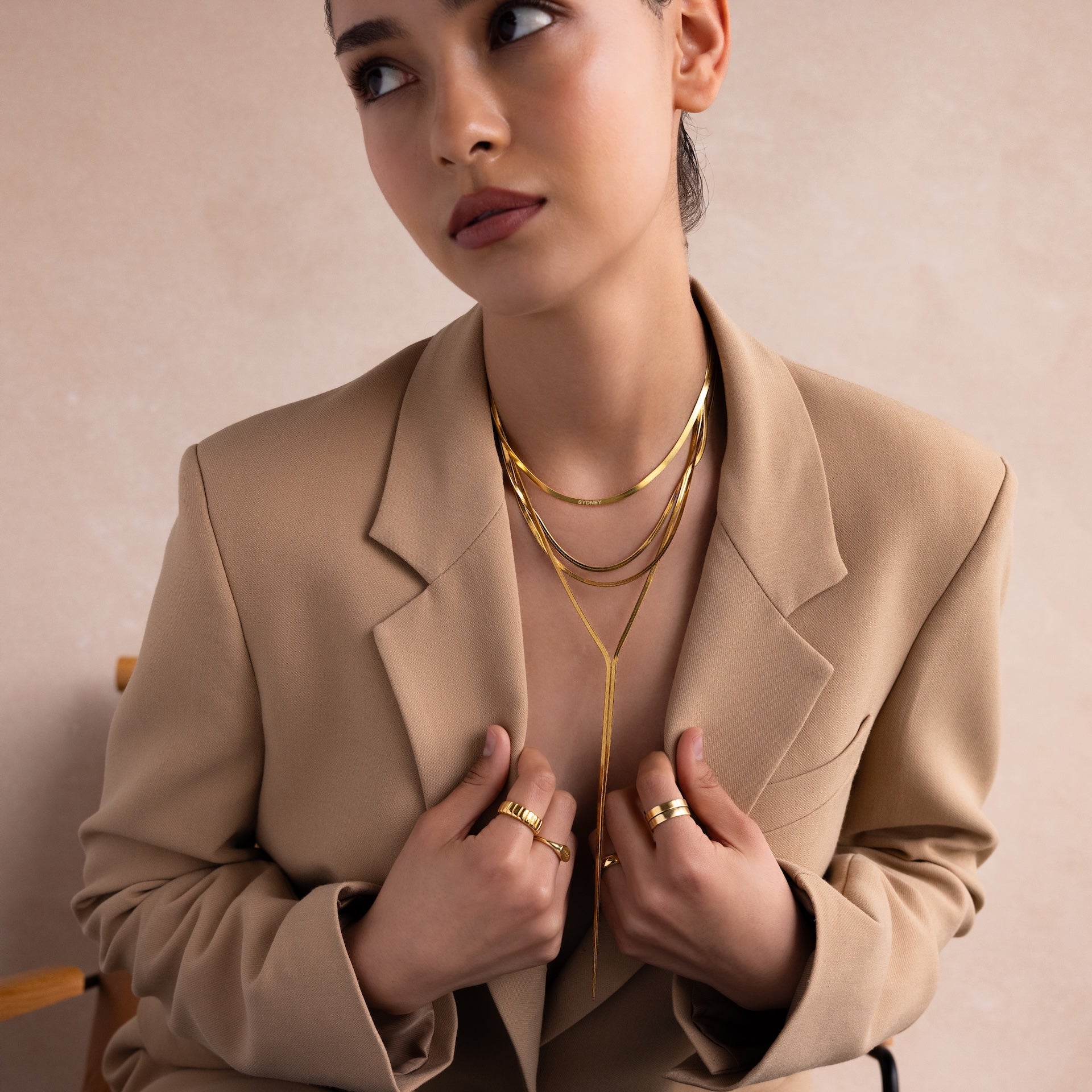 Woman in a tan blazer wearing a layered gold necklace, including the Long Y Herringbone Chain Necklace, looking off to the side.