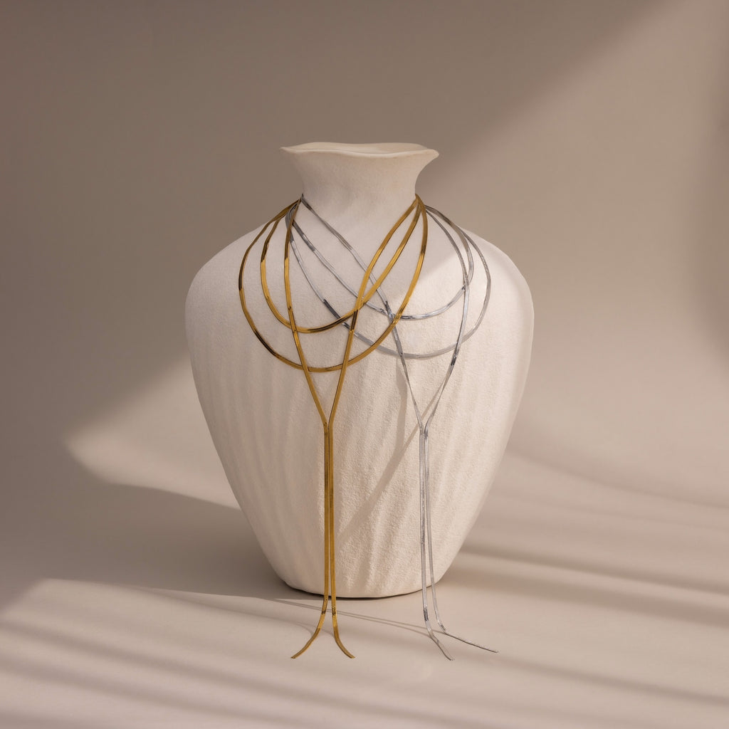 A white textured vase displays the Long Y Herringbone Chain Necklace, draped in layers of gold and silver against a neutral background.