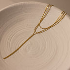 The Long Y Herringbone Chain Necklace, a thin gold-layered piece with multiple strands, rests on a textured white ceramic dish.