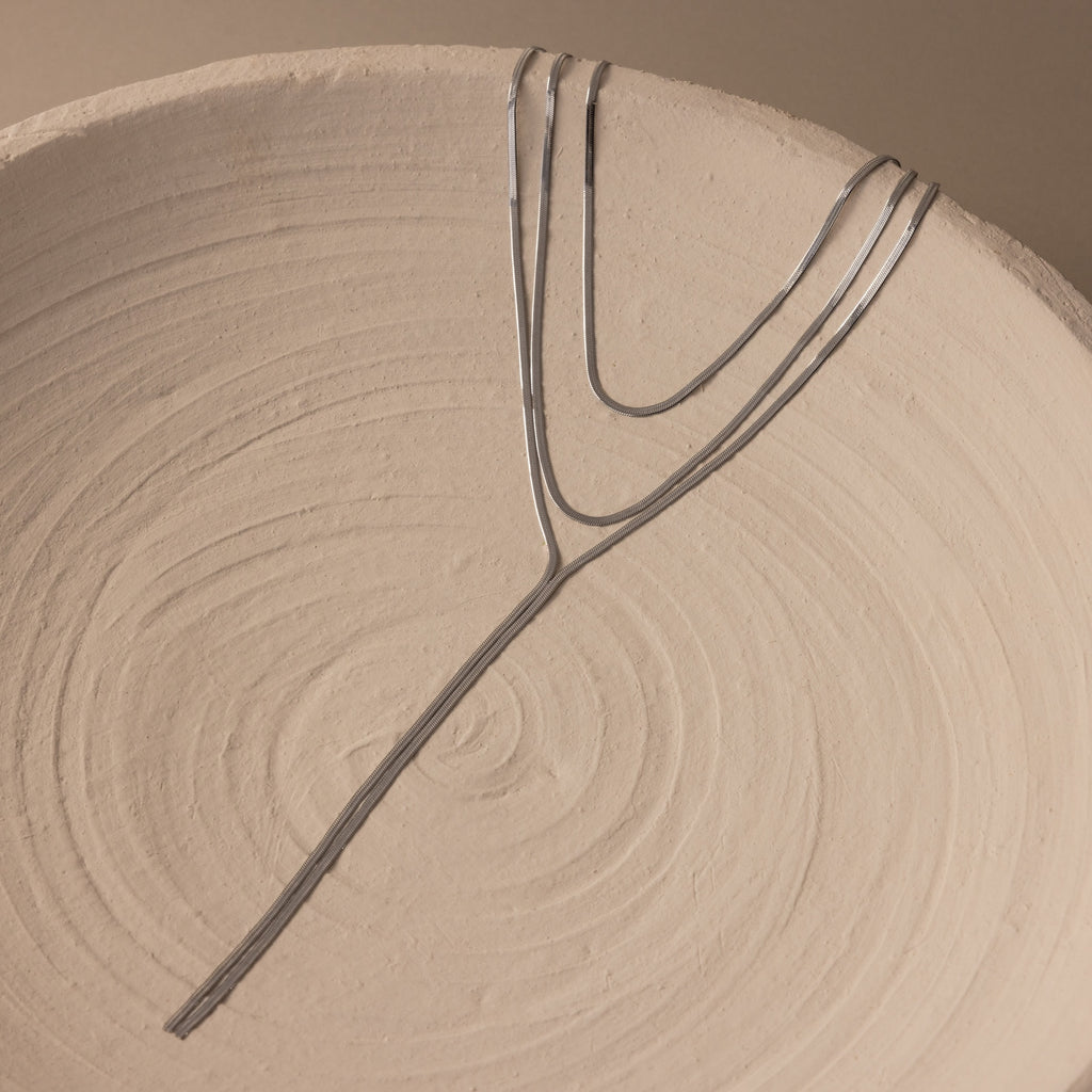 The Long Y Herringbone Chain Necklace, a minimal silver piece, is draped over a textured round ceramic plate with concentric swirl patterns.
