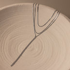 The Long Y Herringbone Chain Necklace, a minimal silver piece, is draped over a textured round ceramic plate with concentric swirl patterns.