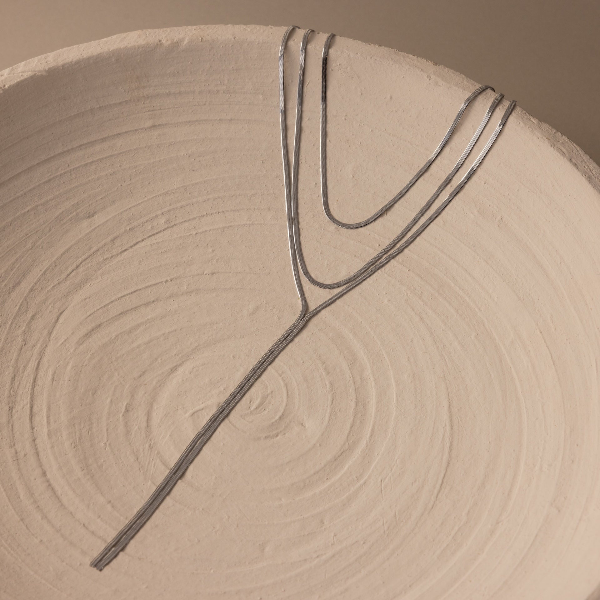The Long Y Herringbone Chain Necklace, a minimal silver piece, is draped over a textured round ceramic plate with concentric swirl patterns.