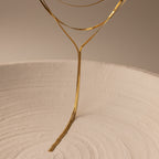 A slender Long Y Herringbone Chain Necklace in 18K Gold elegantly curves above a textured white circular surface, illuminated by soft, warm lighting.