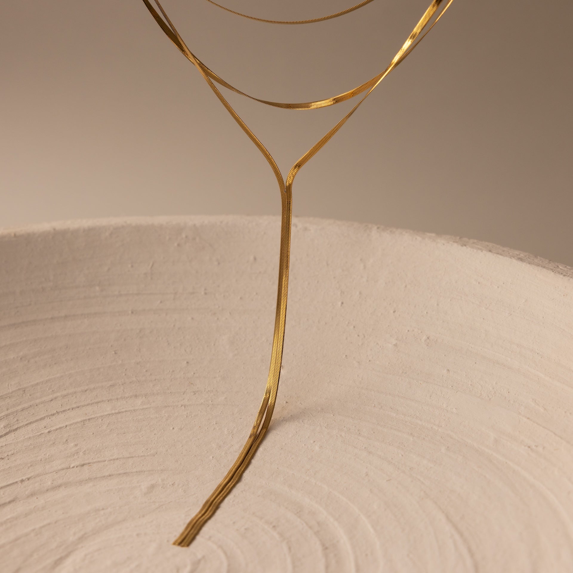 A slender Long Y Herringbone Chain Necklace in 18K Gold elegantly curves above a textured white circular surface, illuminated by soft, warm lighting.