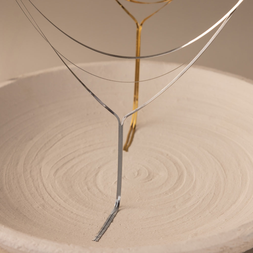 Thin metal wires shaped like trees stand in a textured sand-filled dish, mirroring the refined elegance of the Long Y Herringbone Chain Necklace in Silver.