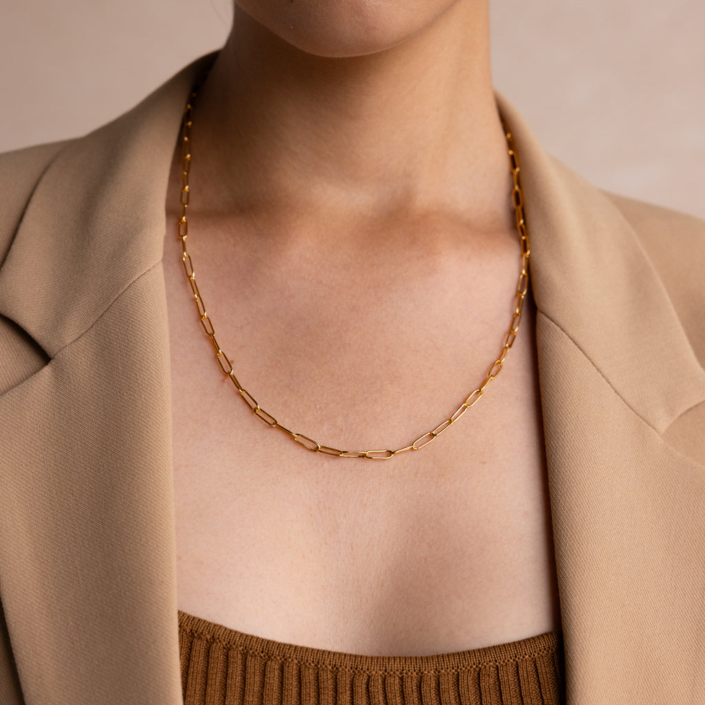 A woman in a beige blazer wears the Paperclip Chain Necklace, a simple gold piece layered over a brown top, showcasing minimalist style.
