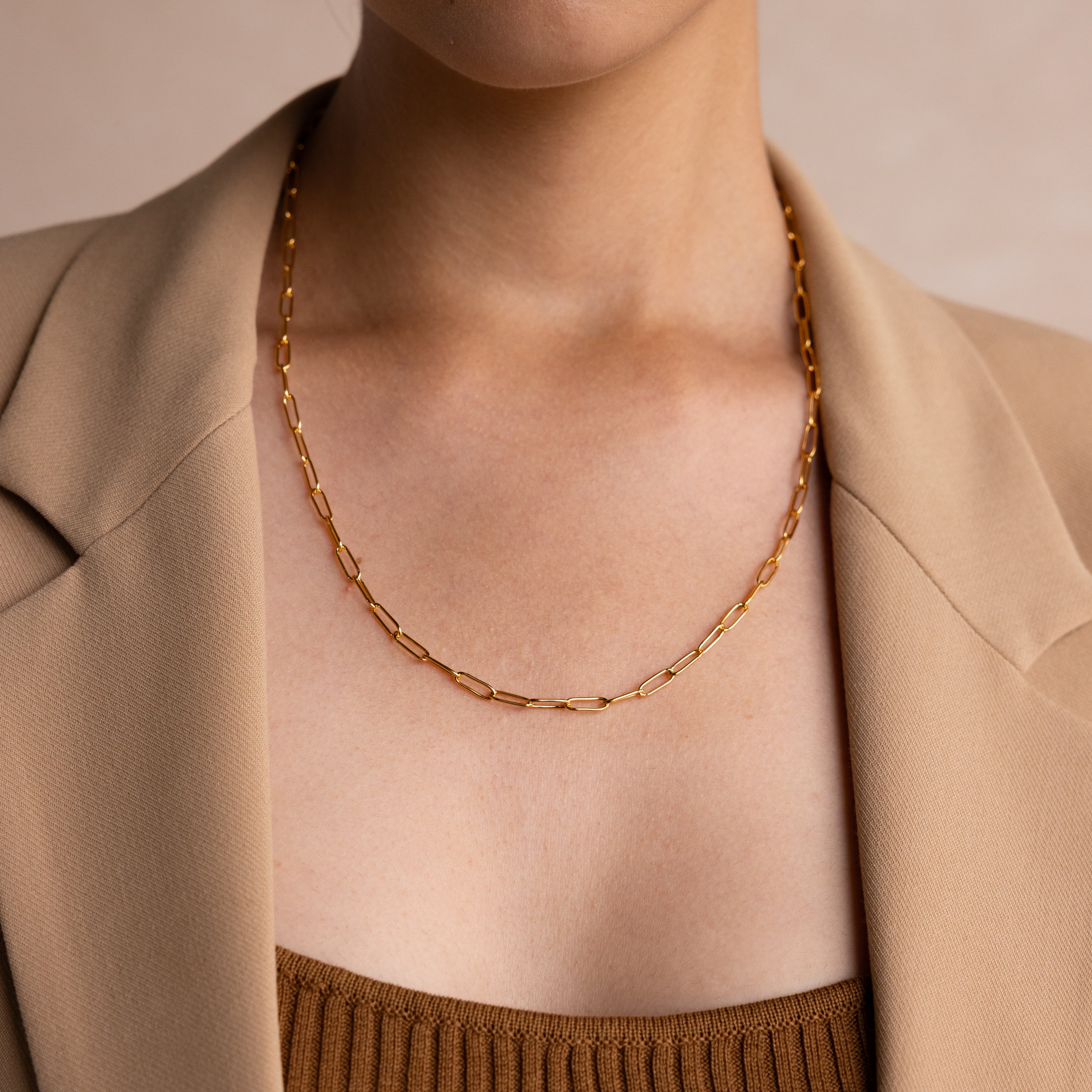 A woman in a beige blazer wears the Paperclip Chain Necklace, a simple gold piece layered over a brown top, showcasing minimalist style.