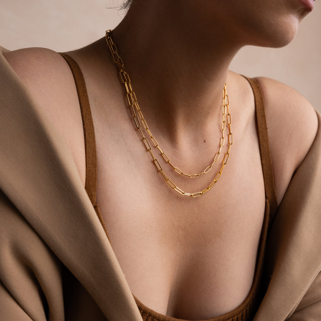 A woman wears the Bold Paperclip Chain Necklace layered with gold necklaces, paired with a brown top and a tan blazer draped over her shoulders.
