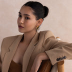 A woman with dark hair in a bun, wearing a tan blazer and gold earrings from our curated jewelry collection, sits on a chair beside our Corporate Gift Box.