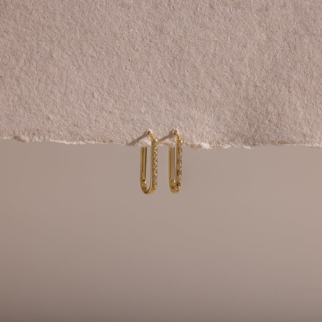 The Dainty Pave Link Earrings, featuring gold hoops with sparkling cubic zirconia stones, hang on a textured, torn beige paper set against a neutral background.
