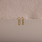 The Dainty Pave Link Earrings, featuring gold hoops with sparkling cubic zirconia stones, hang on a textured, torn beige paper set against a neutral background.