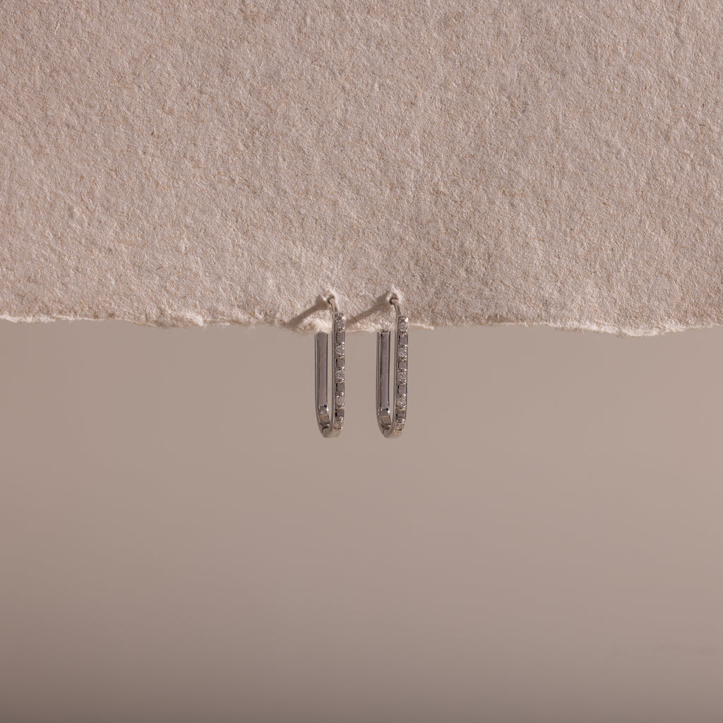 The Dainty Pave Link Earrings, featuring a minimalist silver hoop design, hang from a textured, torn edge of beige paper set against a neutral background.