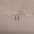 The Dainty Pave Link Earrings, featuring a minimalist silver hoop design, hang from a textured, torn edge of beige paper set against a neutral background.