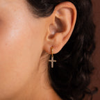 Model wearing gold huggie hoop earrings with dangling pave cross charms.