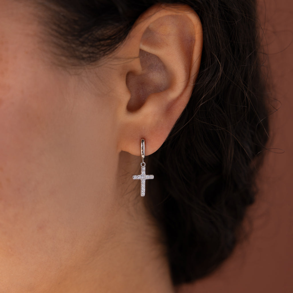 Model wearing silver huggie hoop earrings with dangling pave cross charms.