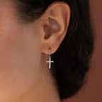 Model wearing silver huggie hoop earrings with dangling pave cross charms.