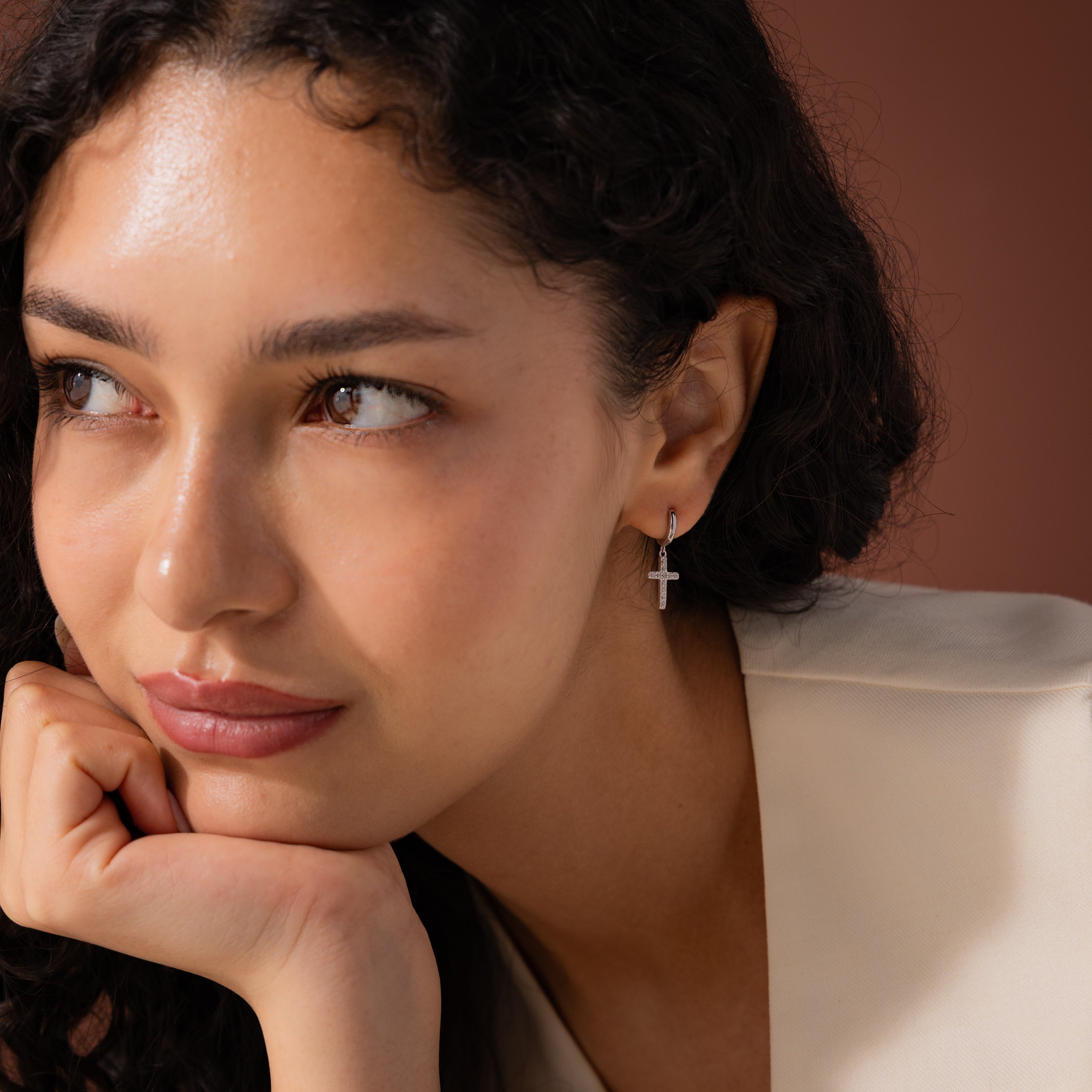 Model wearing silver huggie hoop earrings with dangling pave cross charms.