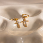 Flat-lay of gold huggie hoop earrings with dangling pave cross charms, displayed on a wavy ceramic surface.