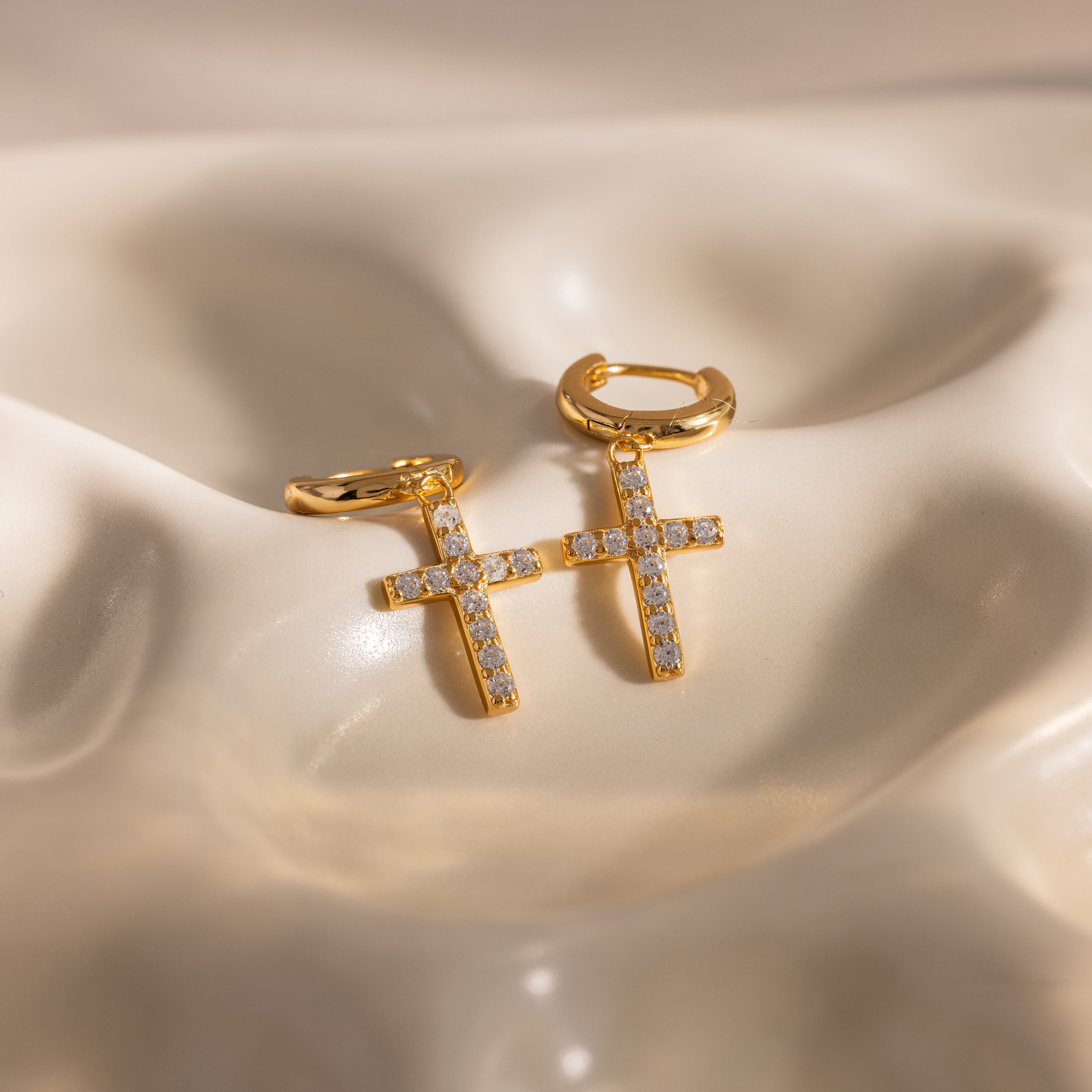 Flat-lay of gold huggie hoop earrings with dangling pave cross charms, displayed on a wavy ceramic surface.